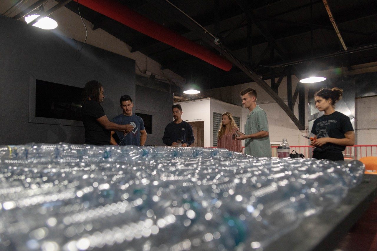 Visiting artist Willie Cole shows Virginia Tech students how to reconfigure plastic bottles for use in a building project. Image credit https://vtnews.vt.edu/
