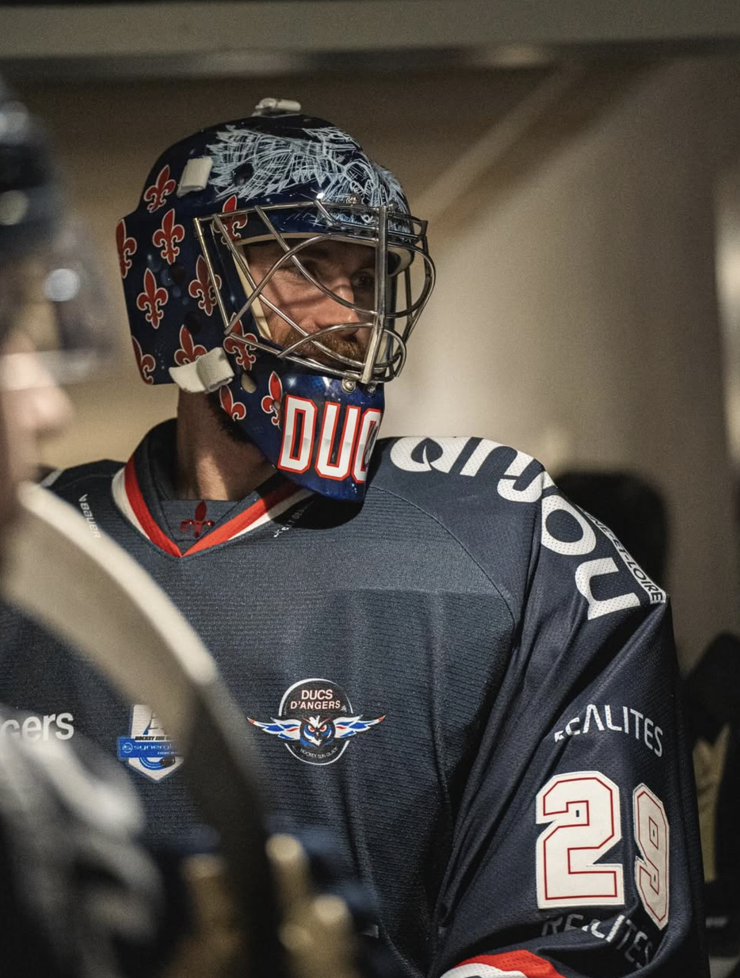 A hockey goalie wearing a black jersey with white, red, and blue accents, a blue helmet with a red and white logo, and a mask with a bird design, smiling through the cage of his helmet.