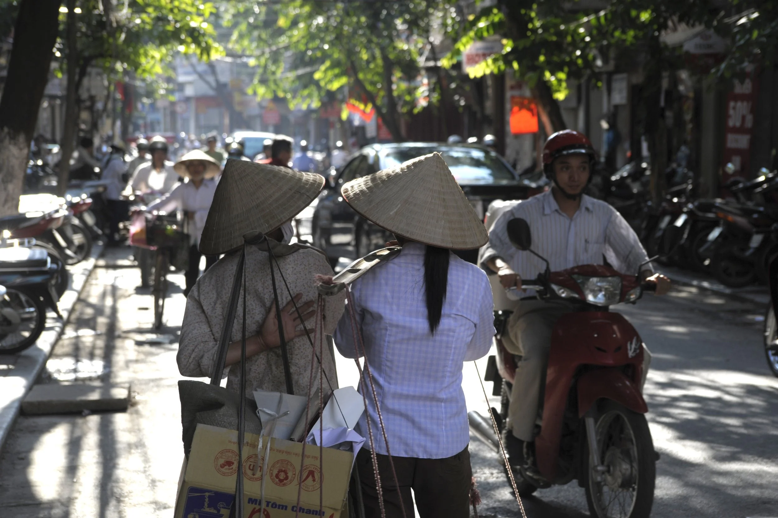 Three women wearing traditional Vietnamese conical hats and carrying shopping bags stand on a busy street, with motorbikes and pedestrians visible in the background.