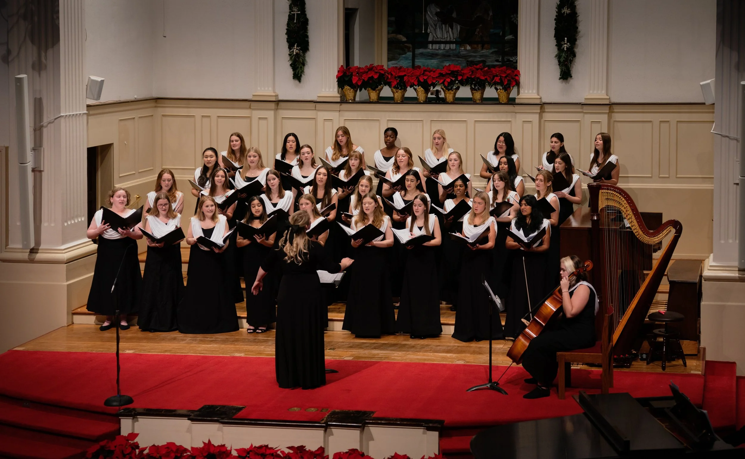 the chorus is performing on stage with a cellist. The singers are wearing black dresses with white drapes and holding music folders. There are red pointsettas above and below.