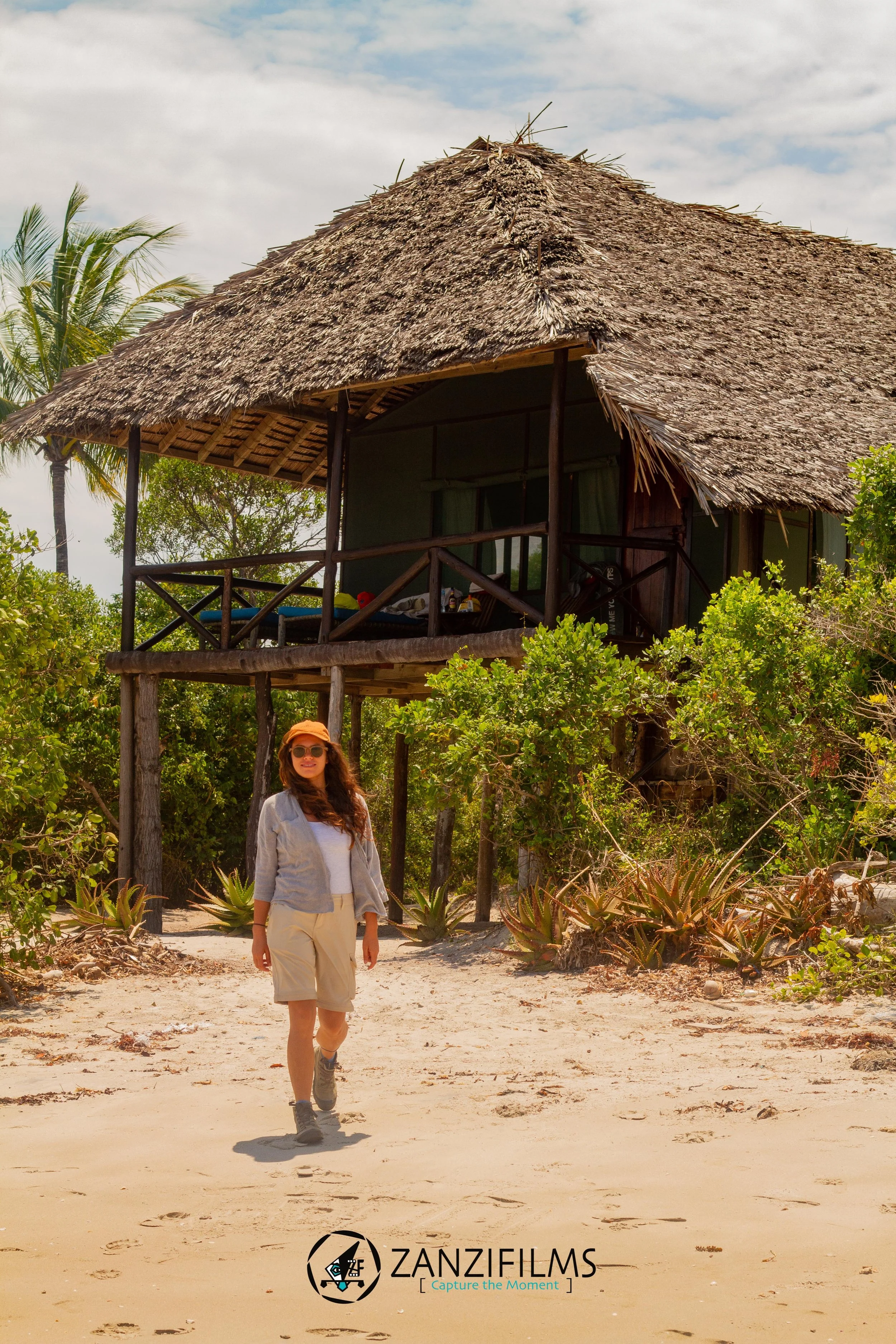 A woman wearing a tan hat, sunglasses, a light gray jacket, and beige shorts walking on a sandy beach with footprints. In the background, there is a wooden hut with a thatched roof, surrounded by green bushes and trees, with a cloudy sky above.