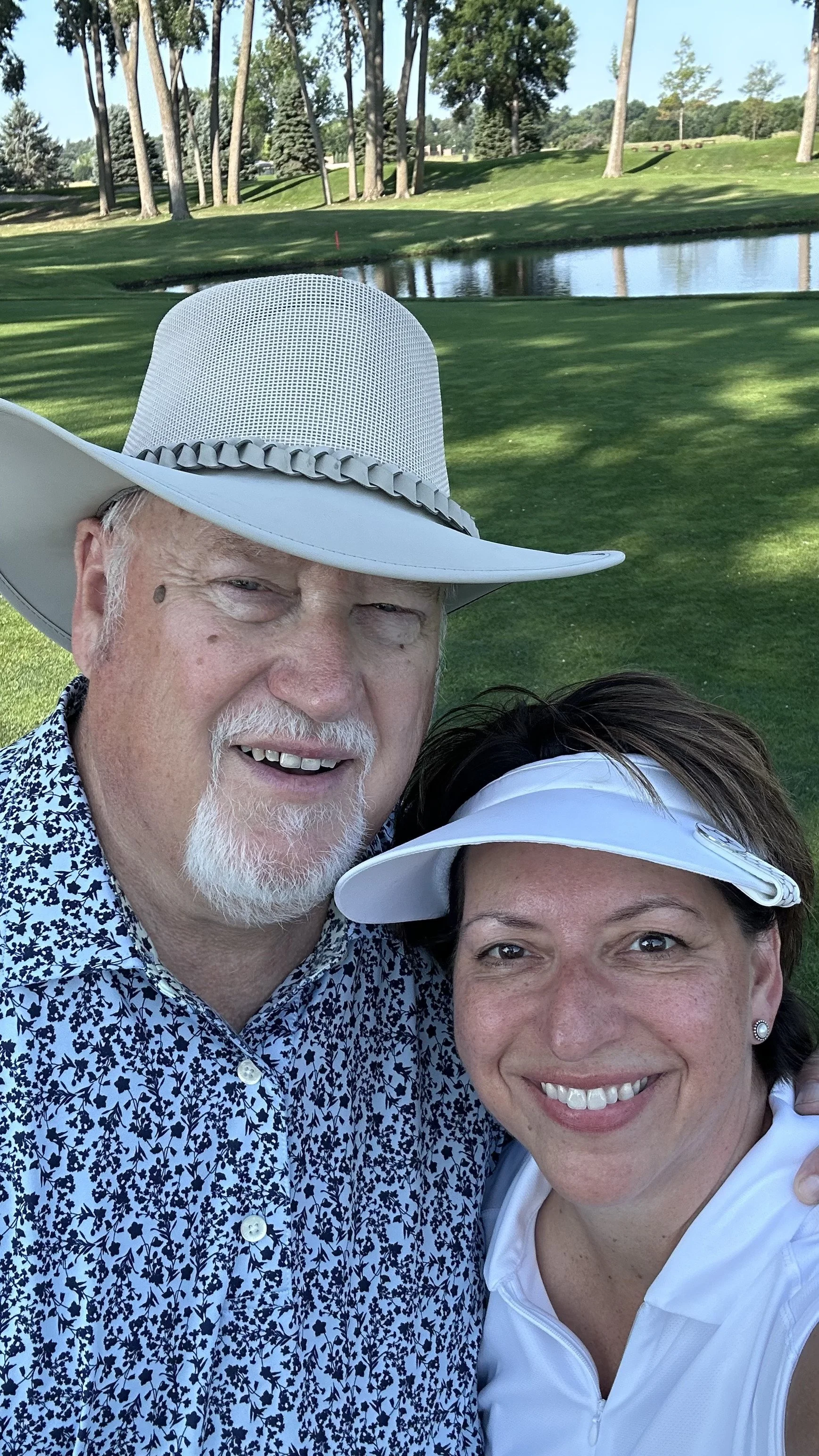 Mike and Anne wearing white sun hats take a selfie beside a pond on a sunny, tree-lined golf course.