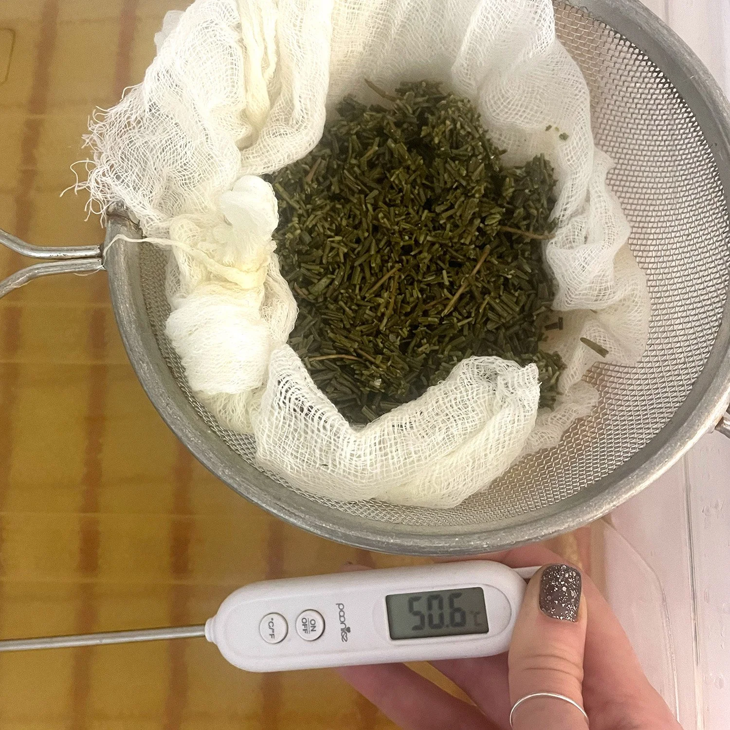 chopped rosemary in muslin cloth, in a sieve over a saucepan - a had is holding a digital thermometer from visual artist Harri Harrison's grief commissions