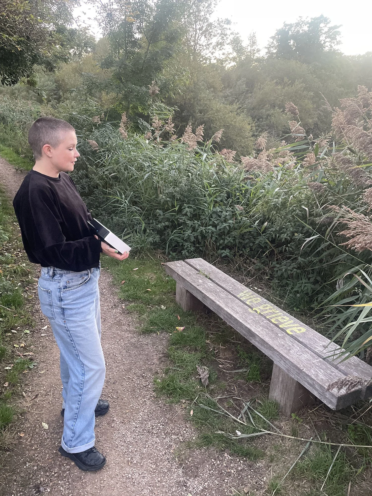 Portrait of Harri Harrison standing outdoors in nature, using a handheld projector to cast text onto a bench