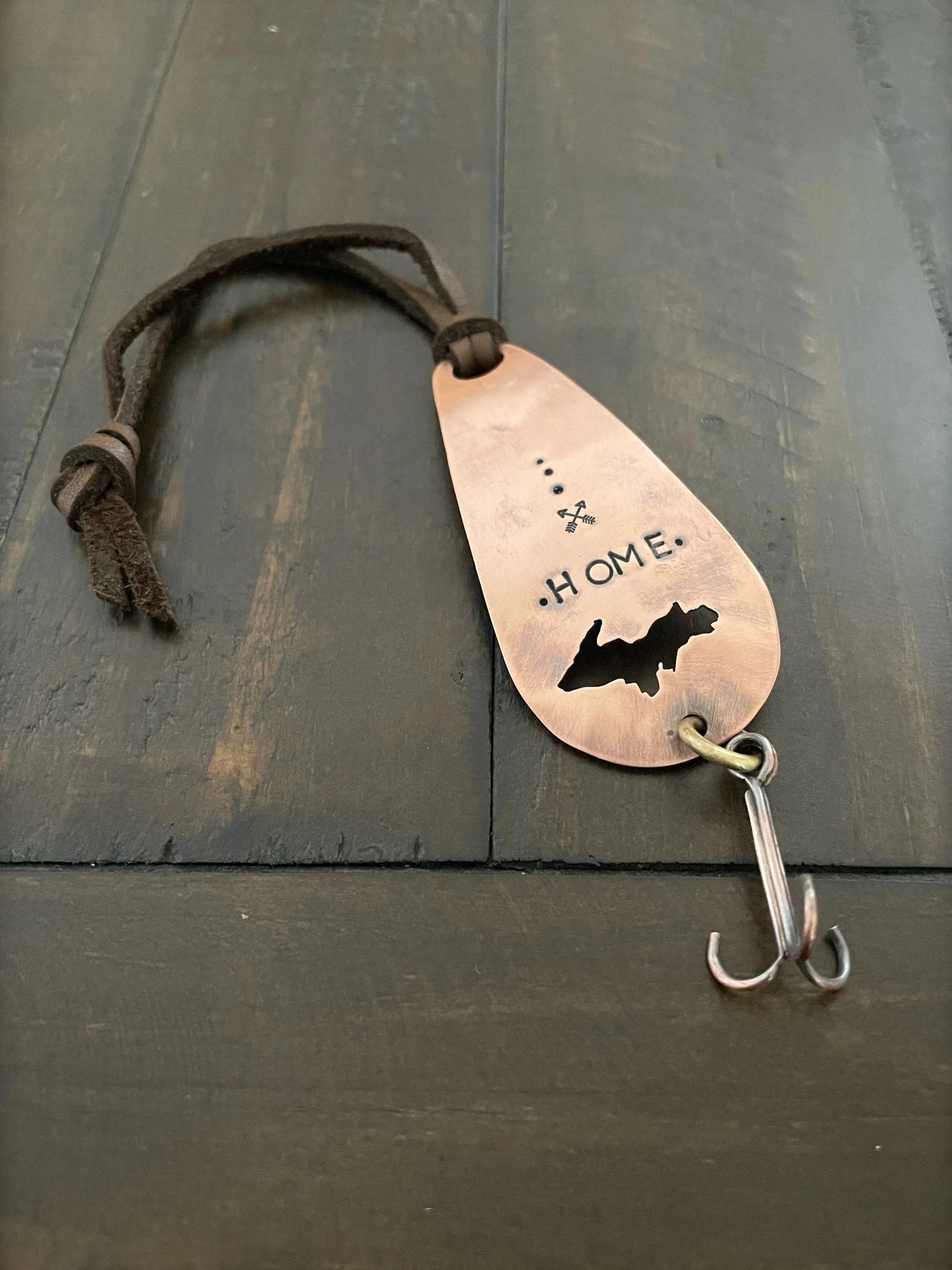 Close-up of a metallic zipper pull attached to a leather strap, resting on a dark wooden surface. The zipper pull has symbols and the word 'HOME' engraved on it, with a silhouette of a house cutout at the bottom.