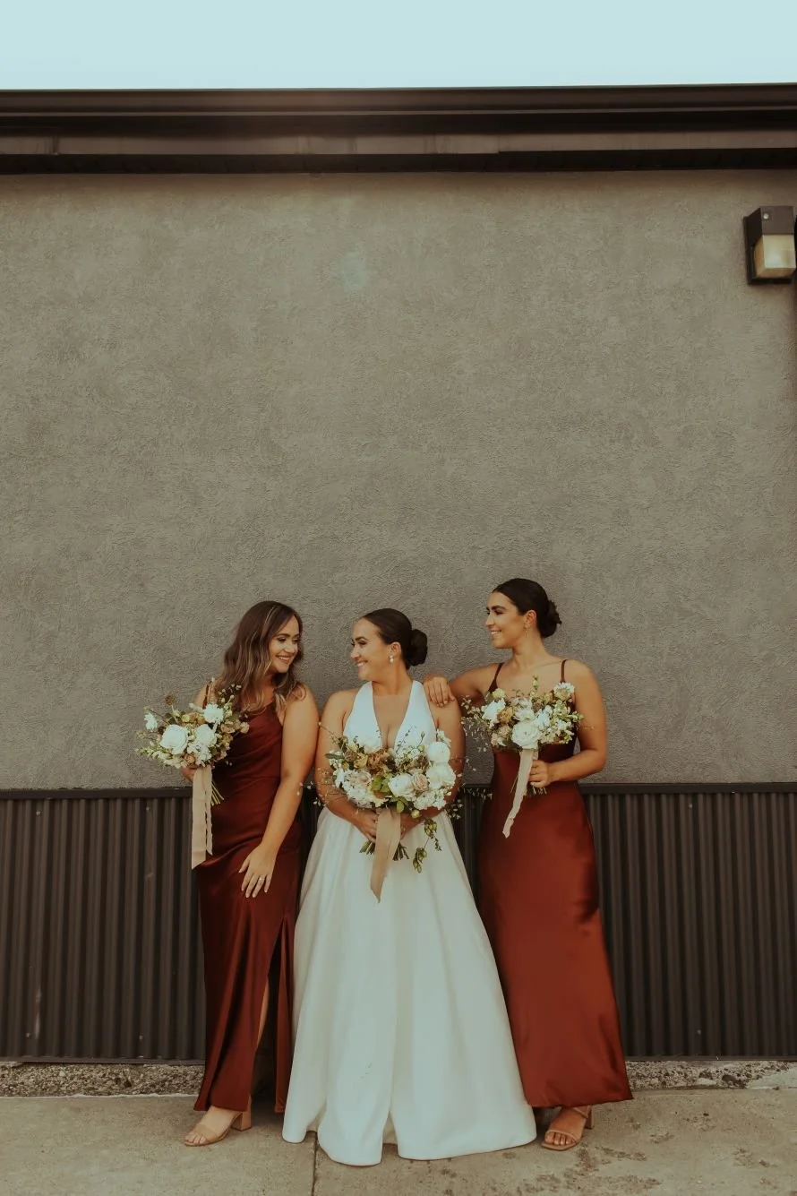 Three women standing against a beige wall, two in maroon dresses and one in a white wedding gown, holding bouquets of flowers, smiling and looking at each other.