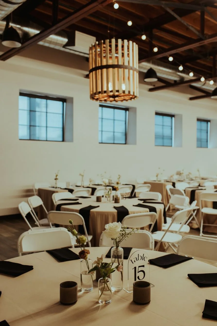 Event space with round tables decorated with black and white table settings, small vases with flowers, and a table sign labeled 'Table 15'. The room has large windows, white walls, a wooden ceiling, and hanging string lights.