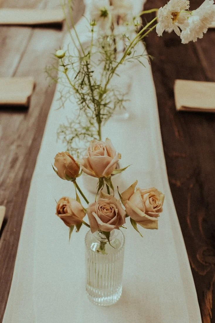 A small glass vase with pale pink roses and greenery on a white table runner, with wooden tables and chairs in the background.