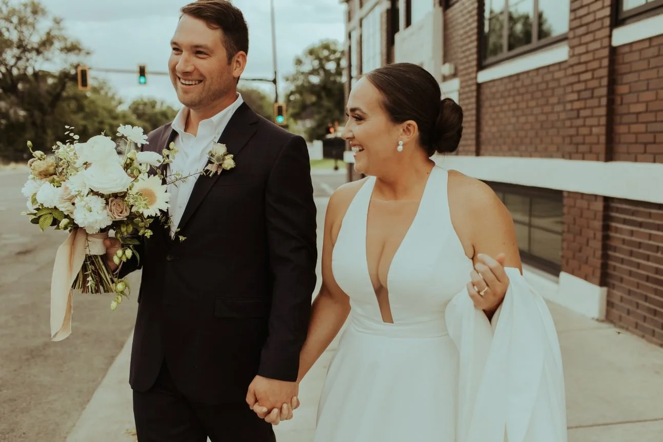 A smiling bride and groom holding hands, walking outdoors on a city street with traffic lights, the bride in a white wedding dress and the groom in a dark suit holding a bouquet of flowers.
