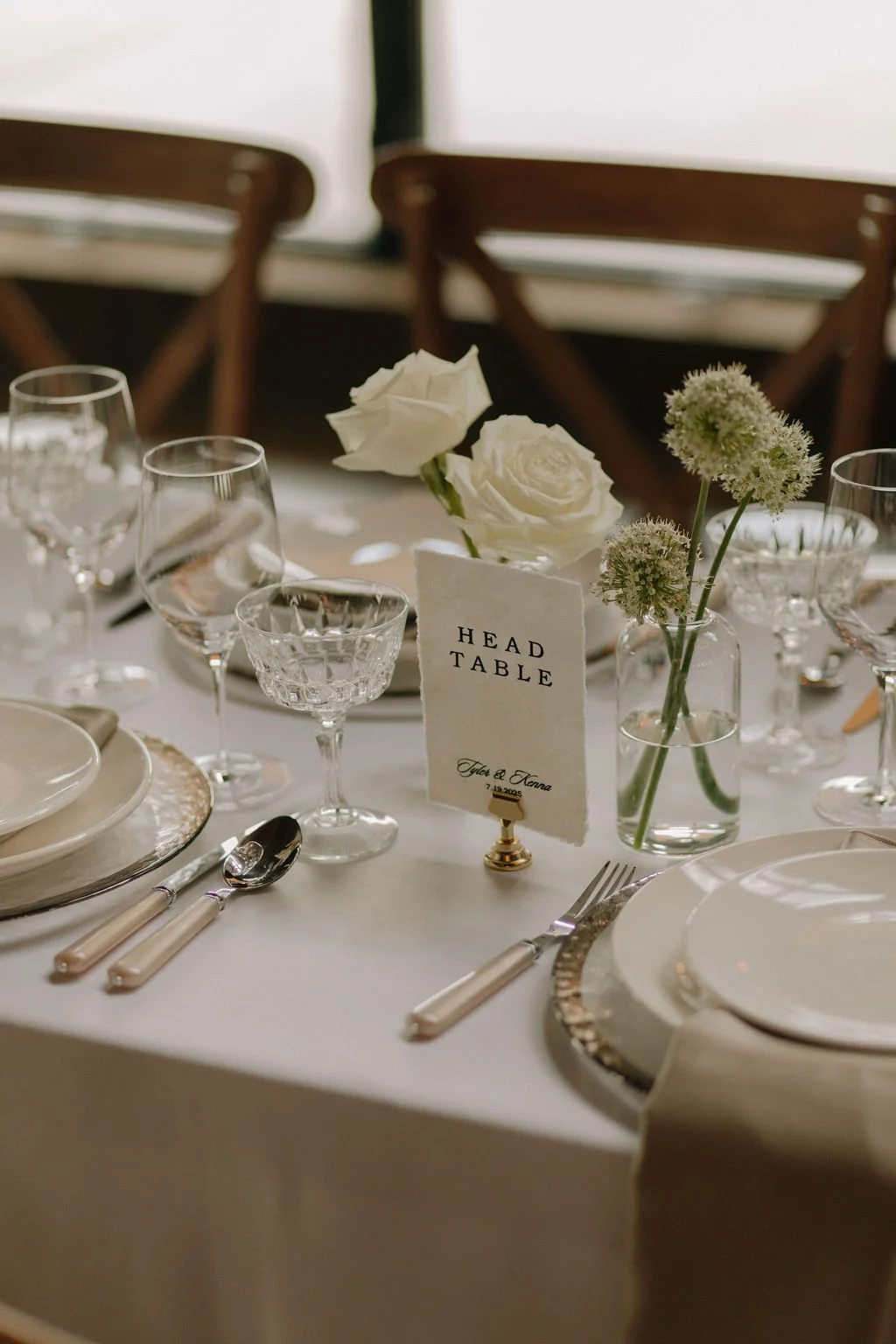 Elegant dinner table setup with white flowers in a vase, glassware, plates, and silverware, with a 'Head Table' sign and place cards.