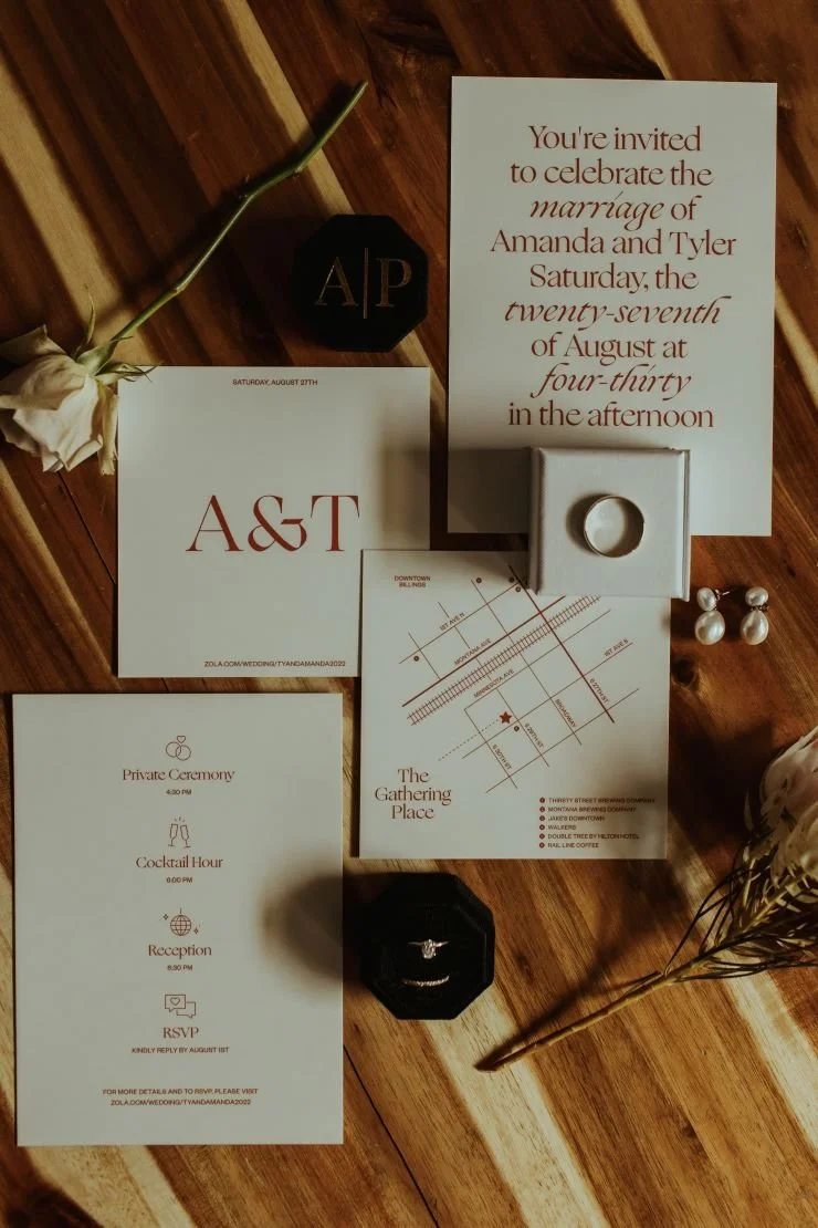 Wedding invitation suite with a white card featuring red text, an envelope with a floral design, black and white jewelry boxes with rings, pearl earrings, a map, and a small pumpkin decor on a wooden surface.
