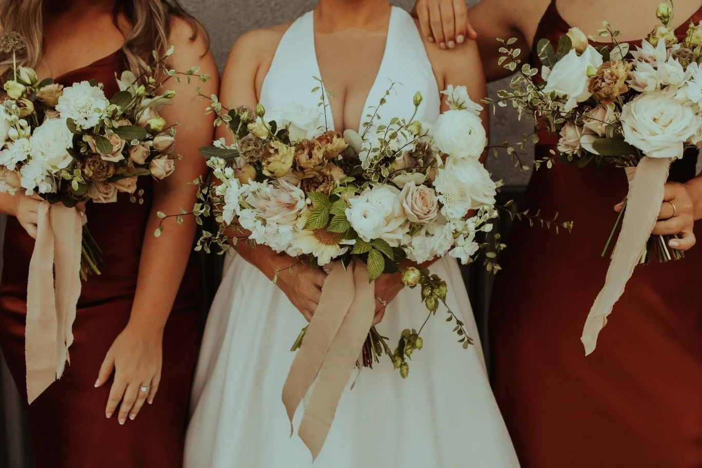 Close-up of a bride and bridesmaids holding bouquets of white and blush flowers with greenery at a wedding.