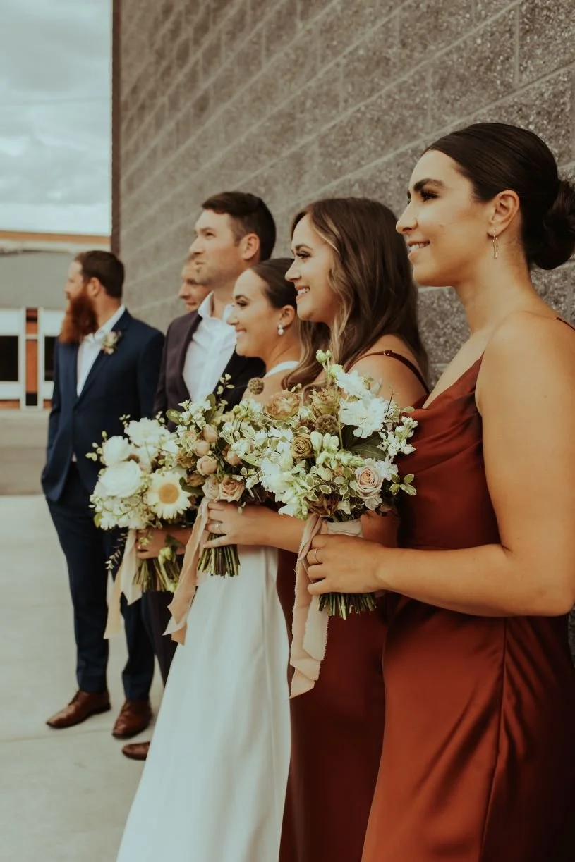 A wedding party standing in a line, each holding a bouquet of flowers, dressed in formal attire, standing against a textured wall.