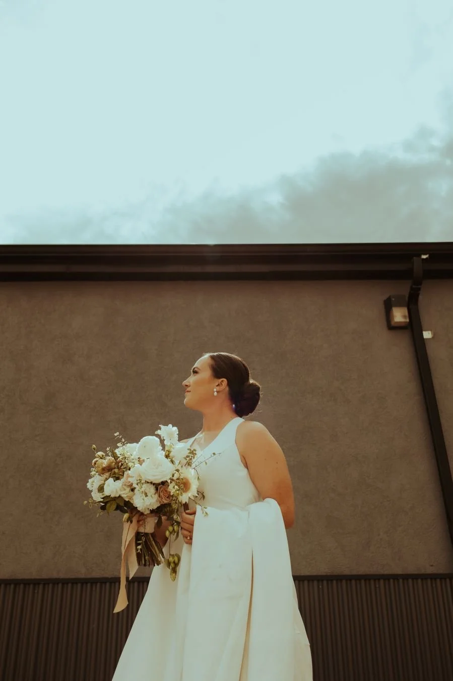 Woman in a white dress holding a bouquet of flowers, standing indoors against a plain wall with an overhead window showing a cloudy sky.