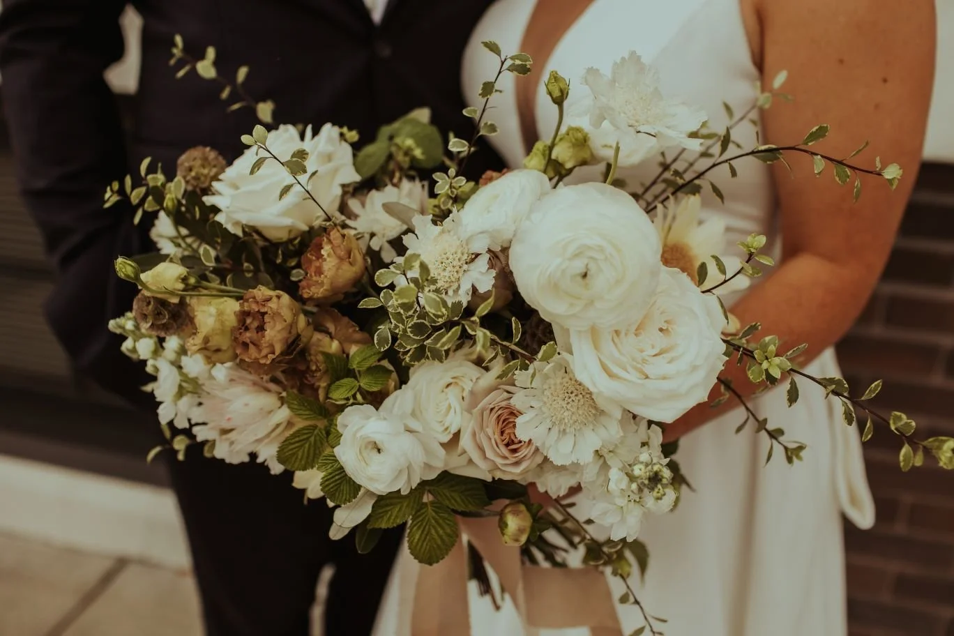 Bride holding a bouquet of white and light pink flowers with greenery, in front of groom wearing a black suit.