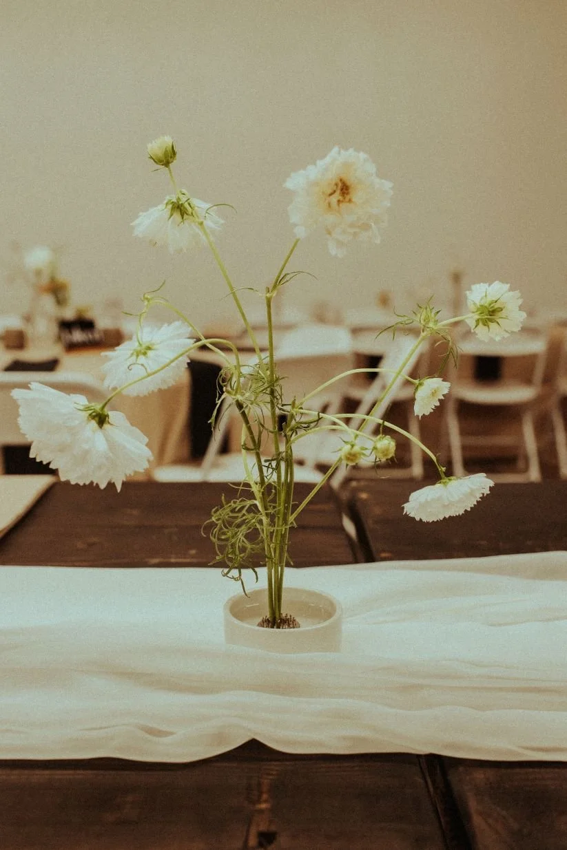 A white flower arrangement in a small vase on a wooden table, with a blurred background of chairs and tables in a dining or event space.