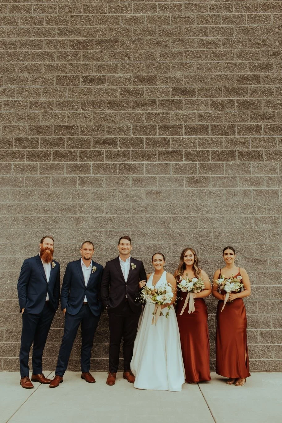 A group of six people standing against a brick wall outdoors, dressed in formal wedding attire with two women holding bouquets.