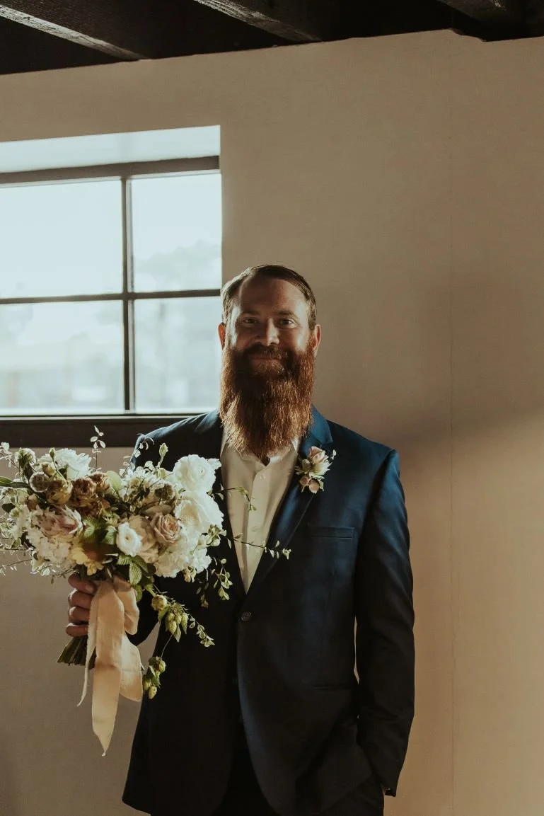 A man with a beard in a dark suit holding a bouquet of white and blush flowers, standing indoors near a window.