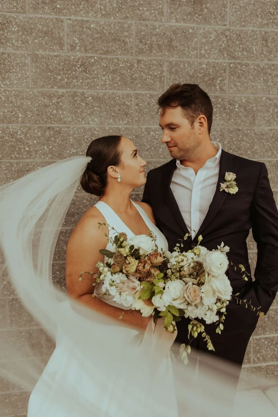 A bride and groom facing each other, holding a bouquet of flowers, standing against a brick wall.