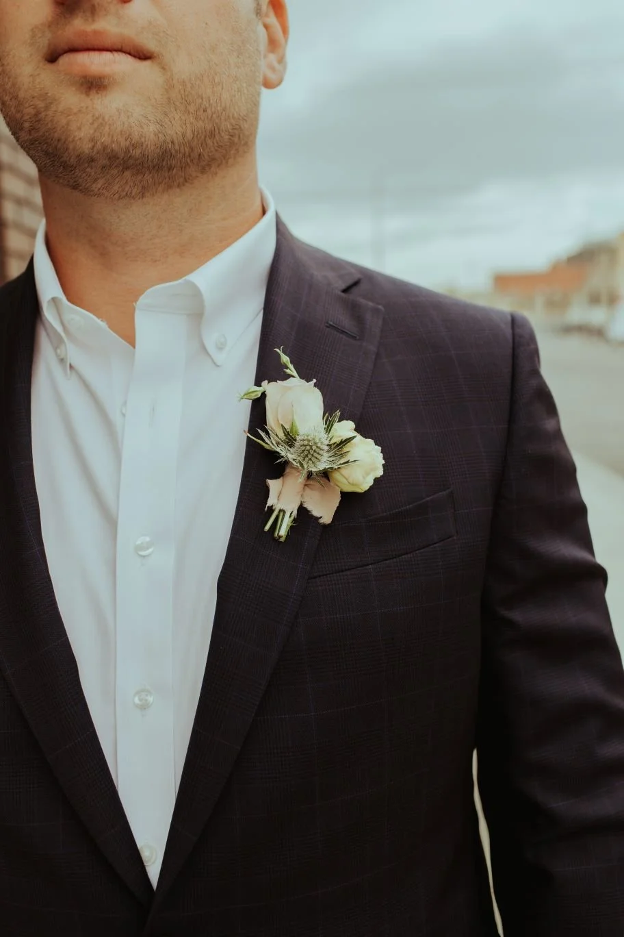 Close-up of a man in a dark suit with a white shirt and boutonniere, standing outdoors on a street.