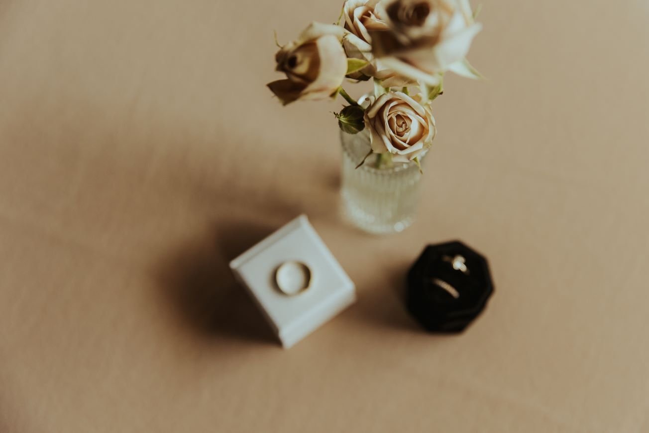 A small glass vase with dried beige roses, a white ring box with a gold ring, and a black velvet ring box with a gold ring inside, set on a beige surface.