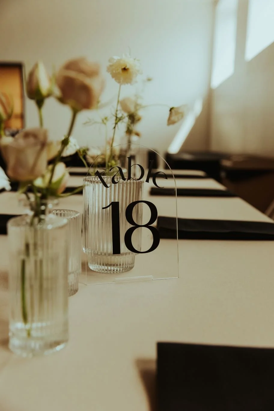 Table setting with a clear acrylic table number sign reading "table 18" in black, surrounded by glass vases with flowers, black napkins, and a beige tablecloth, in a softly lit room.