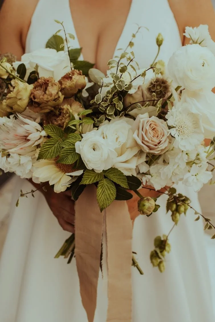 A person holding a large bouquet of white, blush, and brown flowers with greenery, tied with a beige ribbon.