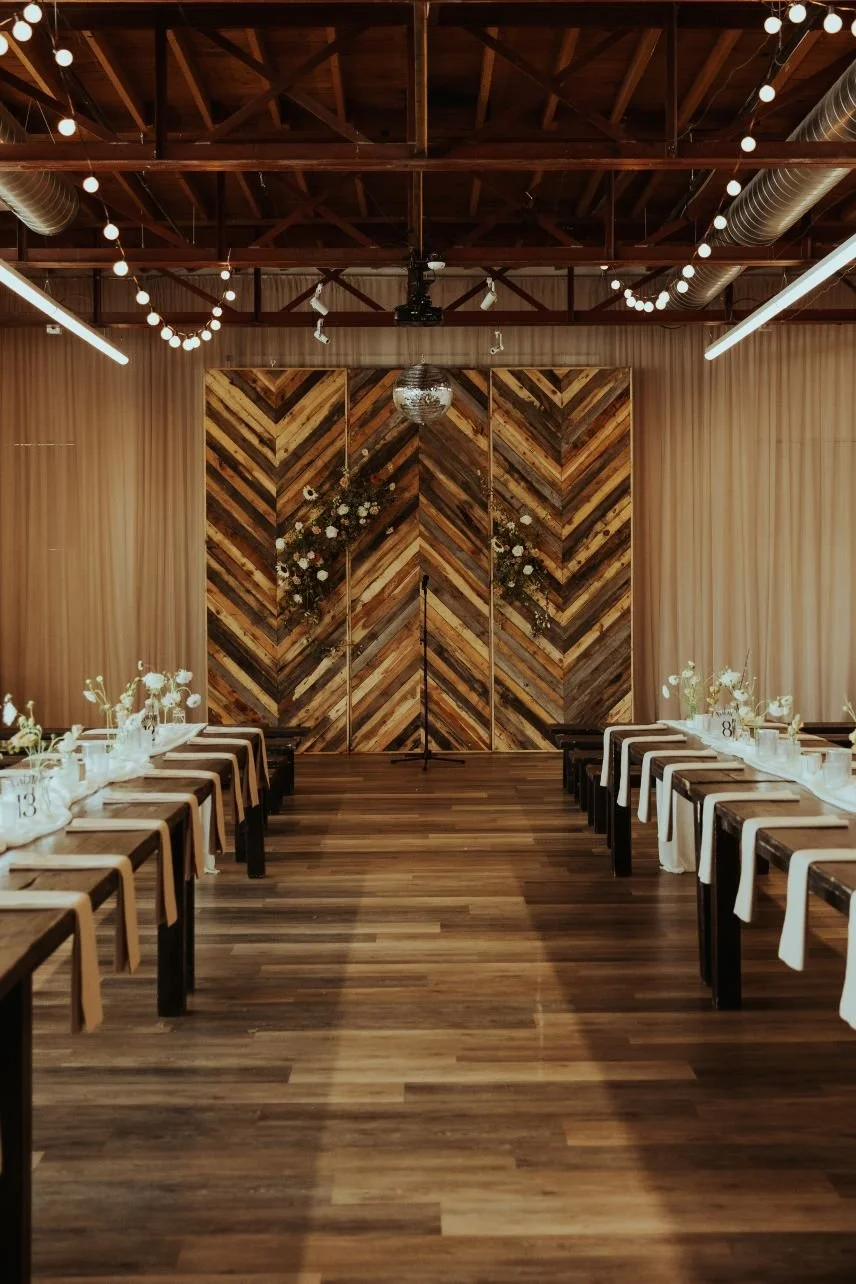 A decorated event space with long tables on either side, adorned with white tablecloths, flowers, and table numbers. A wooden chevron-patterned wall with hanging floral arrangements is at the front, with string lights hanging from the ceiling.