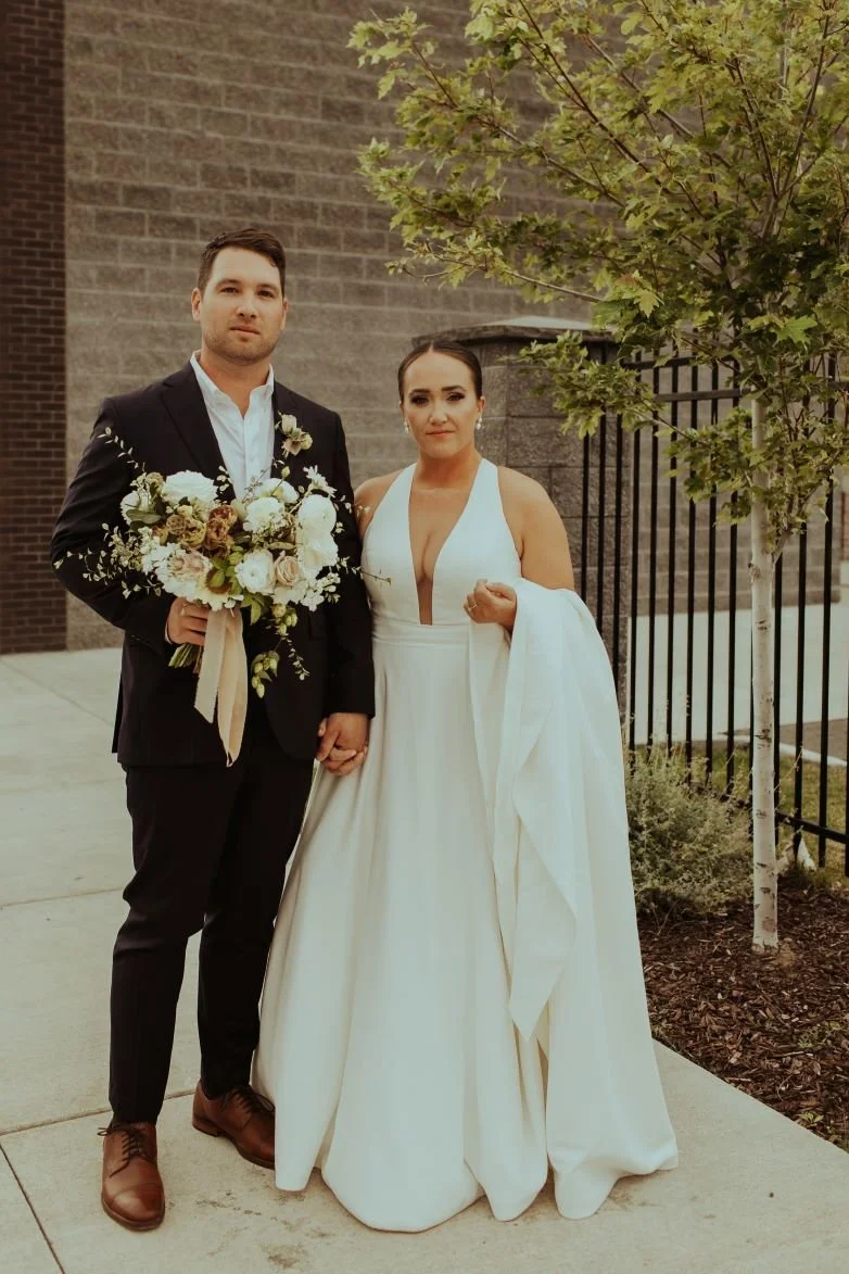 A bride and groom standing outdoors, holding hands. The bride is wearing a white gown with a deep neckline, and the groom is in a black suit with a white shirt. The groom is holding a bouquet of white and cream flowers. There is a small tree and a br