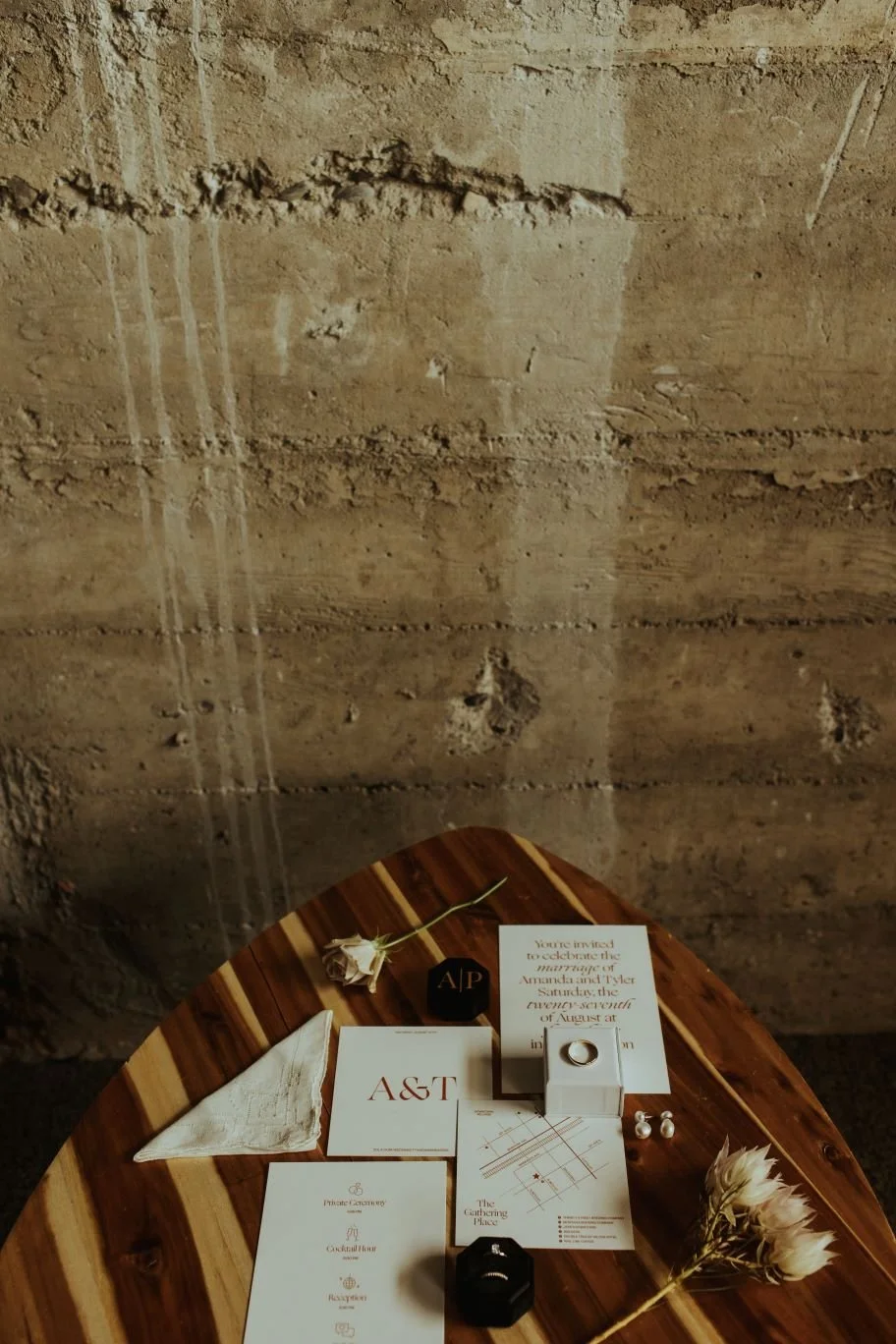 Wedding invitation and details, boutonniere, and decorative items on a wooden table against a textured brick wall.
