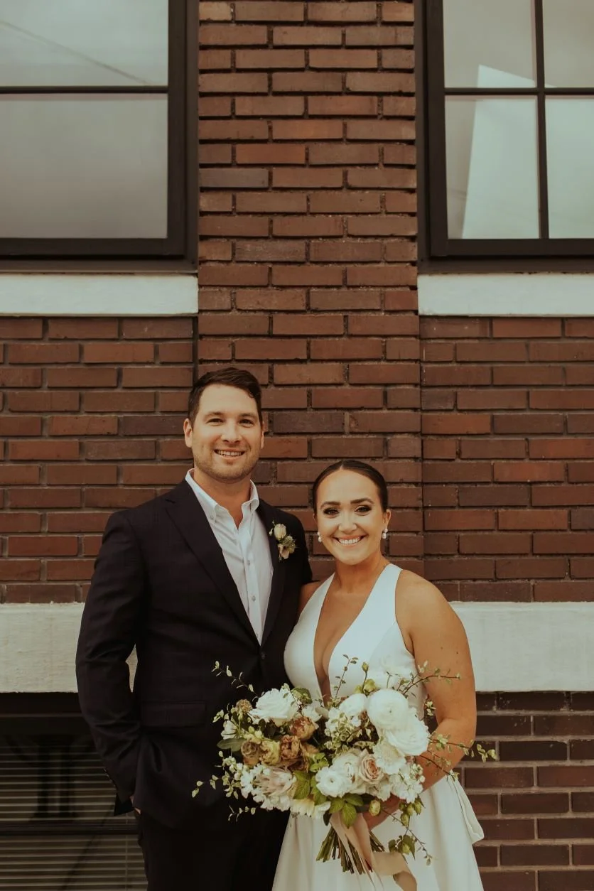 A newlywed couple standing in front of a brick wall, smiling, with the bride holding a large bouquet of white and blush flowers. The groom is dressed in a black suit with a white shirt, and the bride is wearing a white wedding gown with a deep V neck