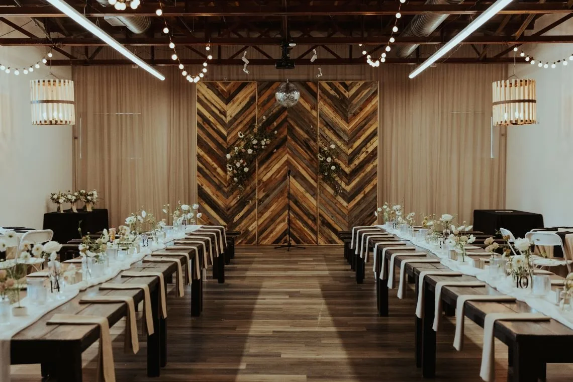 Elegant event space with long wooden tables decorated with white flowers in glass jars, set with napkins and cutlery, and framed by beige curtains, string lights, and a wooden chevron-patterned backdrop at the front.