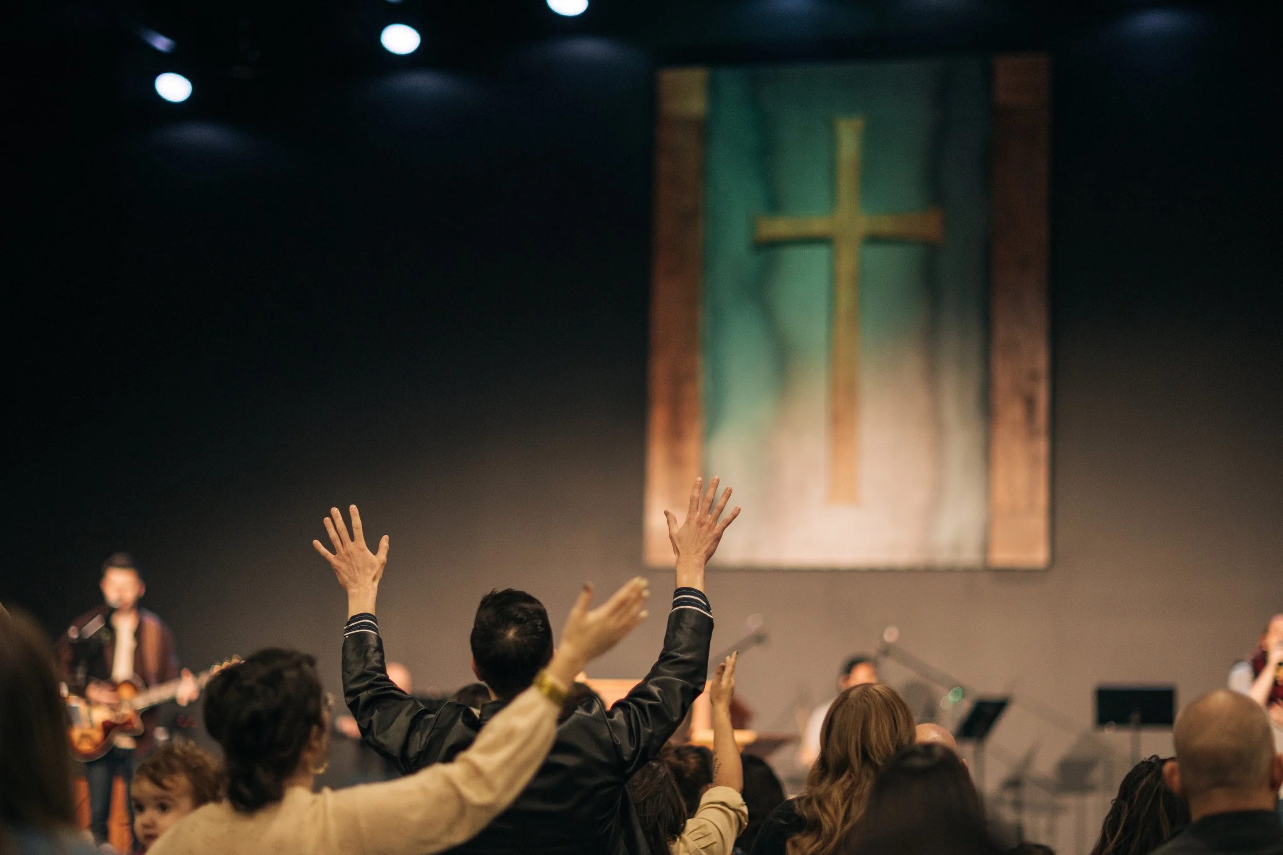 People raising their hands in a church congregation during worship with a cross on a banner hanging behind the stage.