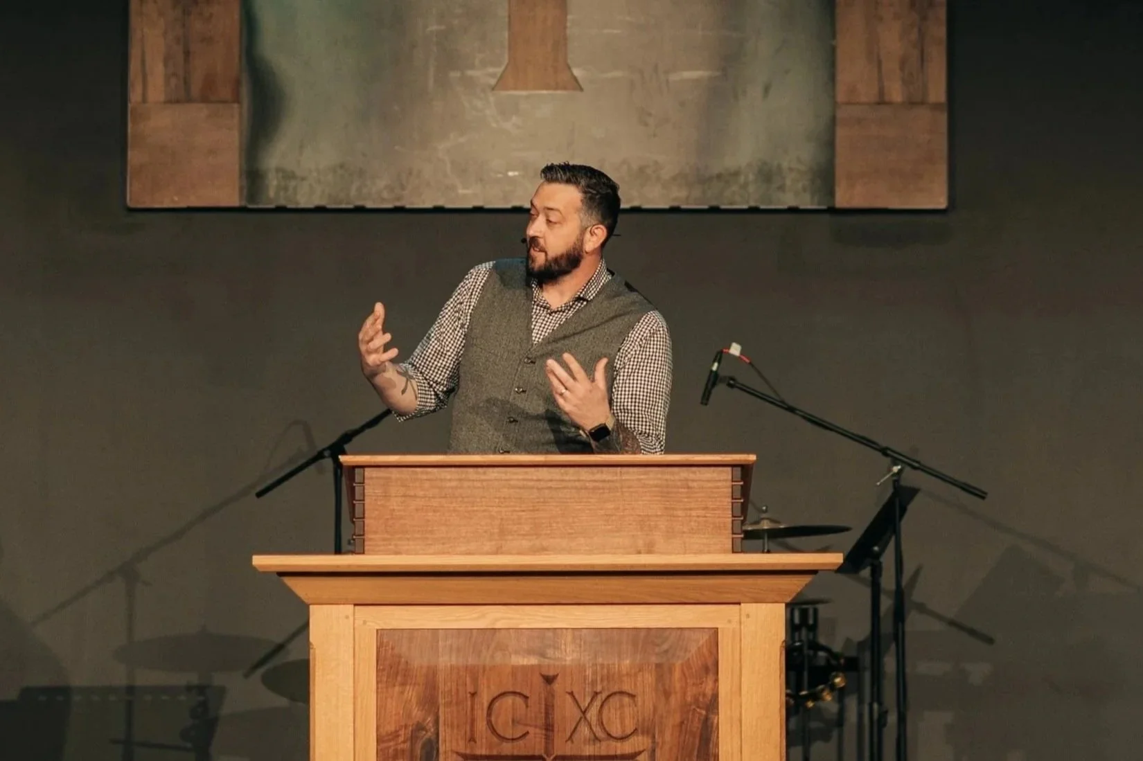 A man standing behind a wooden podium, speaking with expressive hand gestures during a presentation or speech in an indoor setting.