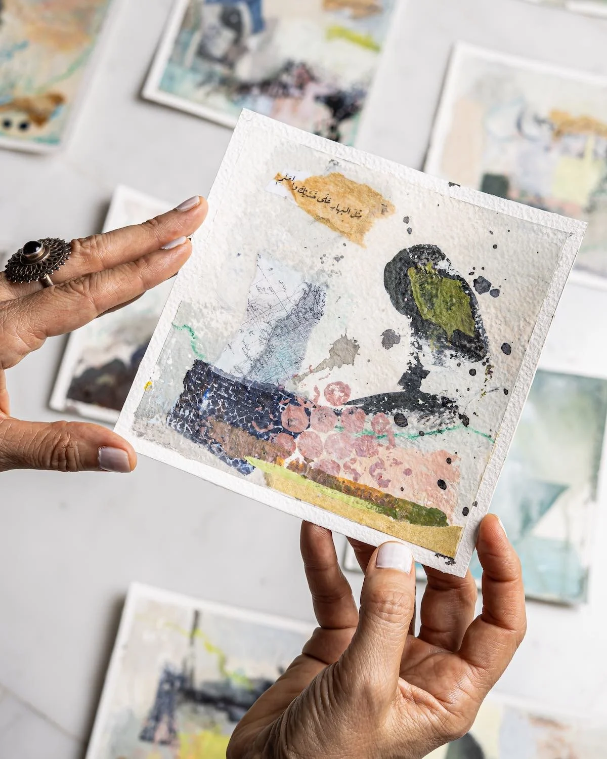 Pale as Bahar, a 15 × 15 cm painting from the Burda Collection, held in Sara Kabariti's hands above the rest of the collection laid out on marble