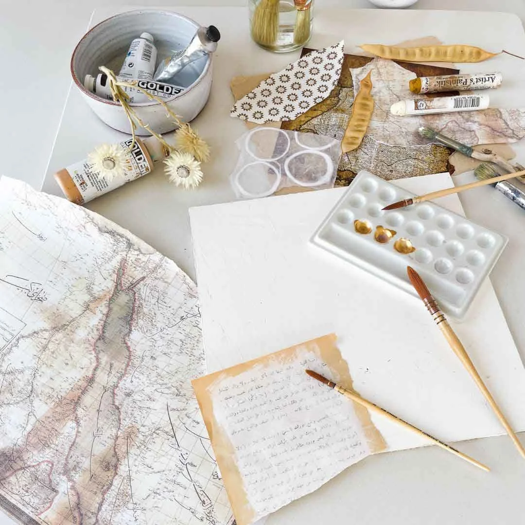 Studio materials for the Hu Collection - a tube of gold paint, paint sticks, tubes of paint in a white ceramic bowl, dried flowers, vintage map fragments, a floral patterned paper and a sheet of handwritten Arabic text, by Sara Kabariti