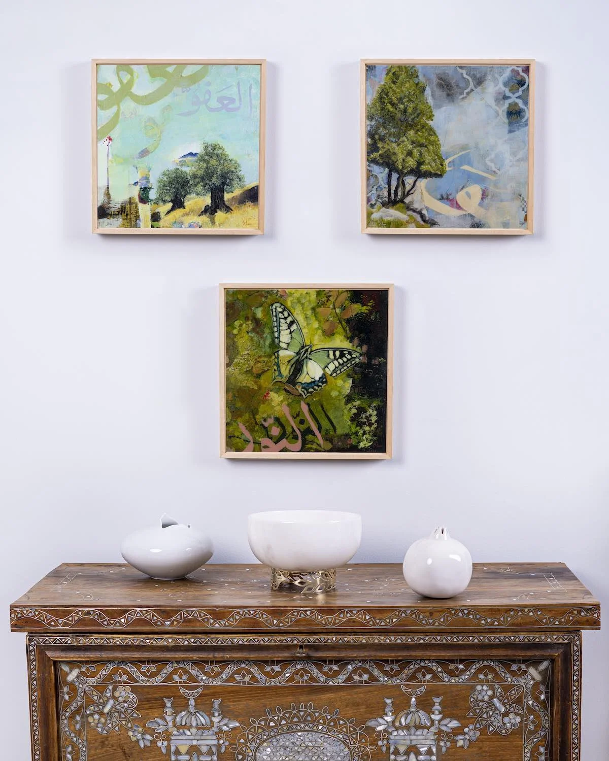Unveiling Truth and Balmy Meadow partially visible, with New Beginnings shown in full, framed together from the Hu Collection above a carved wooden console table with white ceramic vessels, mixed media on panel by Sara Kabariti