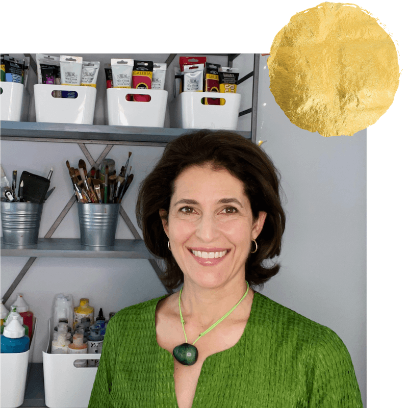 Sara Kabariti smiling in her studio, wearing a green top and a green necklace, standing in front of shelves holding paint supplies, brushes and collage materials