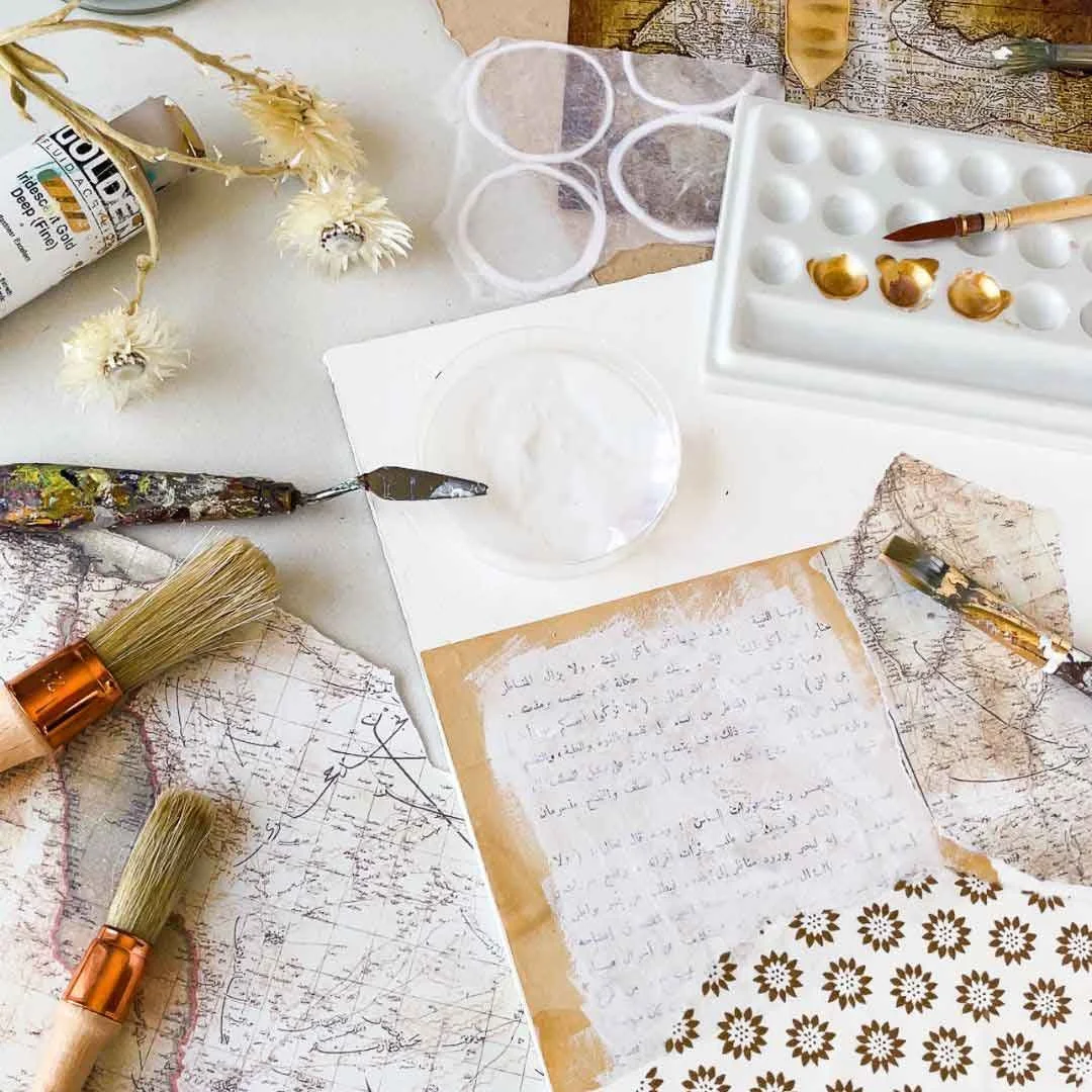 Studio materials for the Hu Collection - gold paint, a palette knife with white paint, paintbrushes, dried flowers, vintage map fragments, a floral patterned paper and a sheet of handwritten Arabic text, by Sara Kabariti