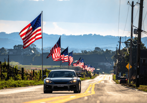 Car driving on a rural road lined with American flags, with rolling hills and utility poles in the background.