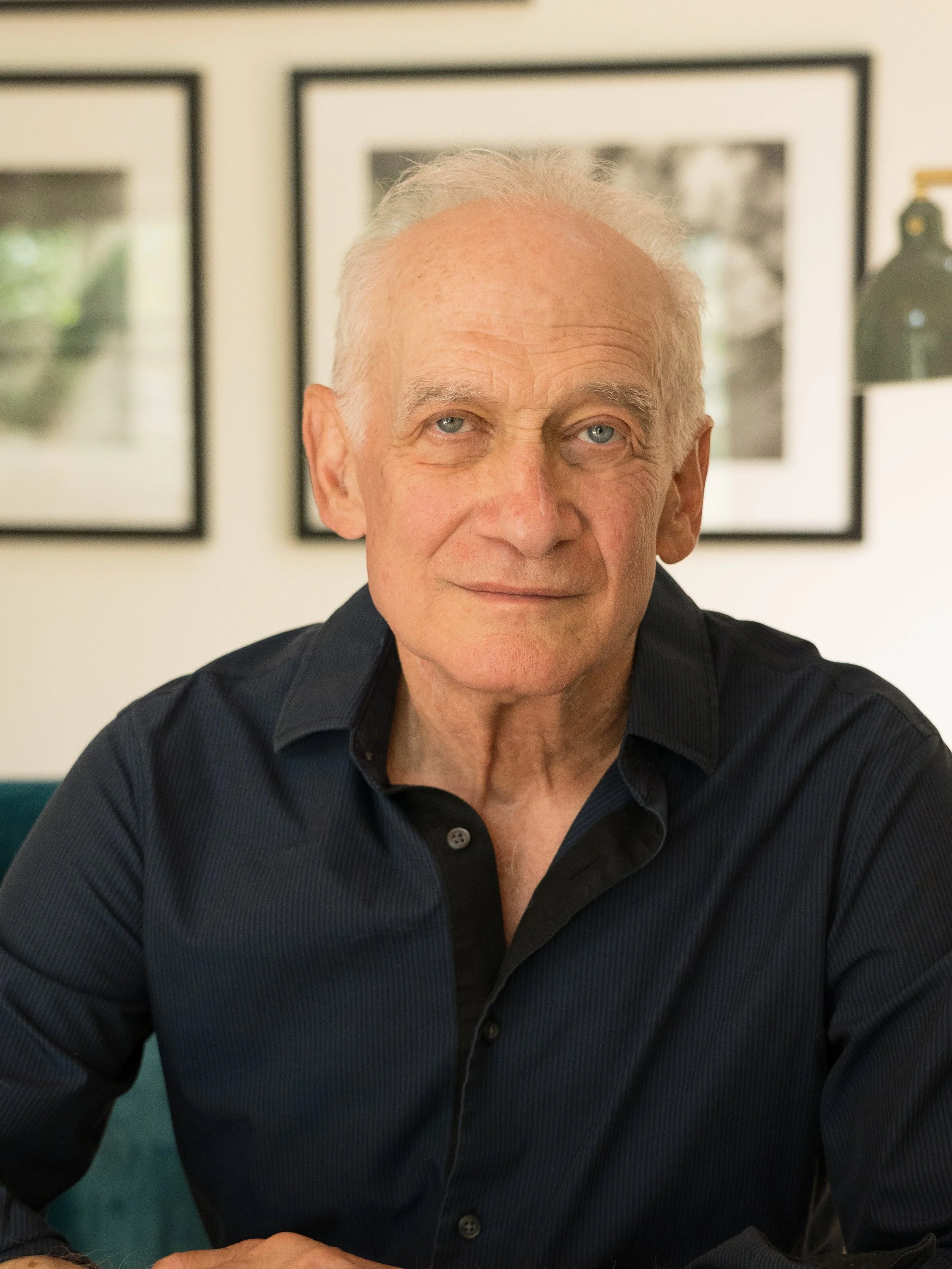 An elderly man with light skin and short gray hair, wearing a dark button-up shirt, sitting indoors with framed photos on the wall behind him.