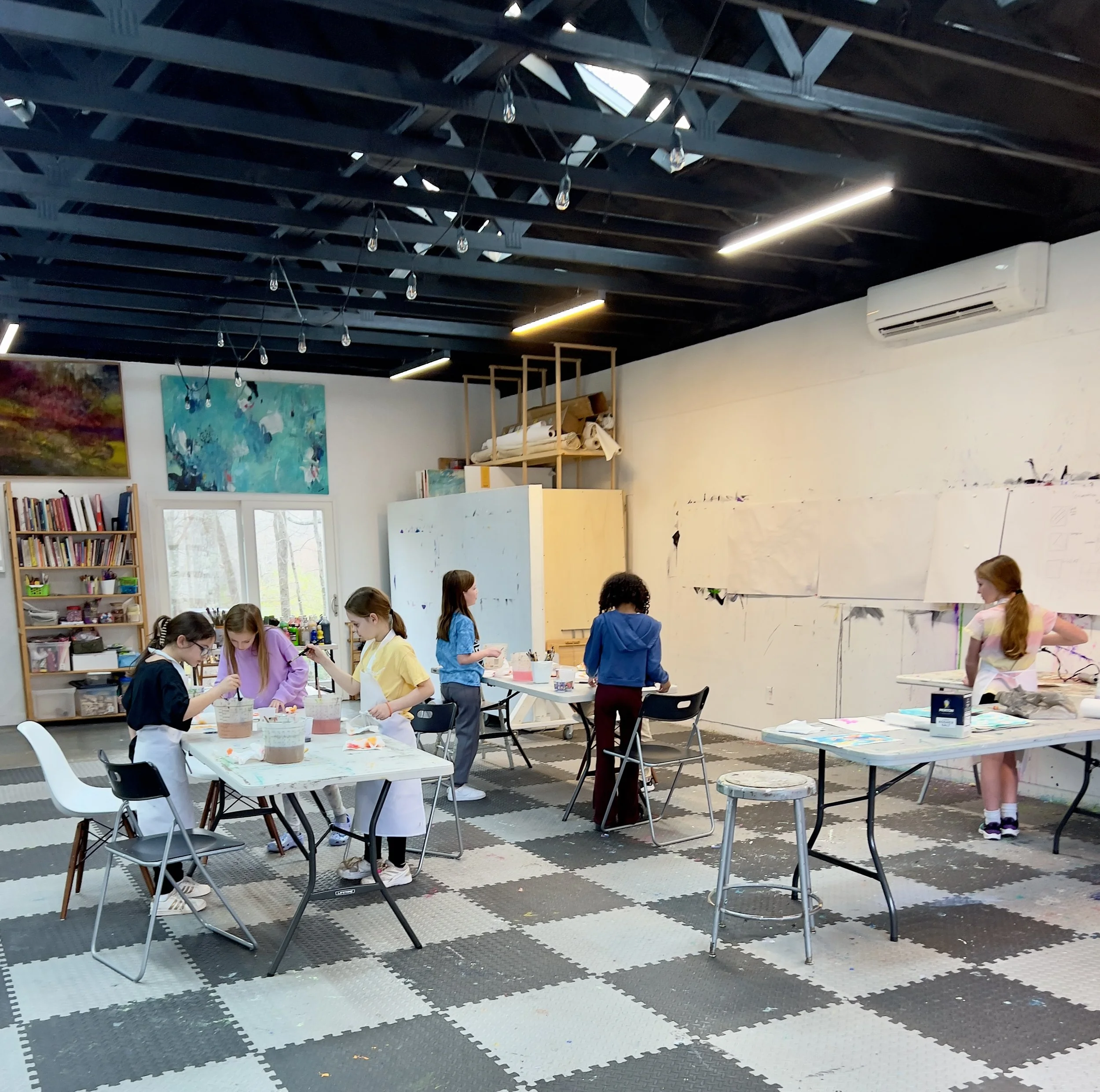 Children in an art classroom painting and drawing at tables with art supplies, colorful paintings on the wall, and a checkered floor.