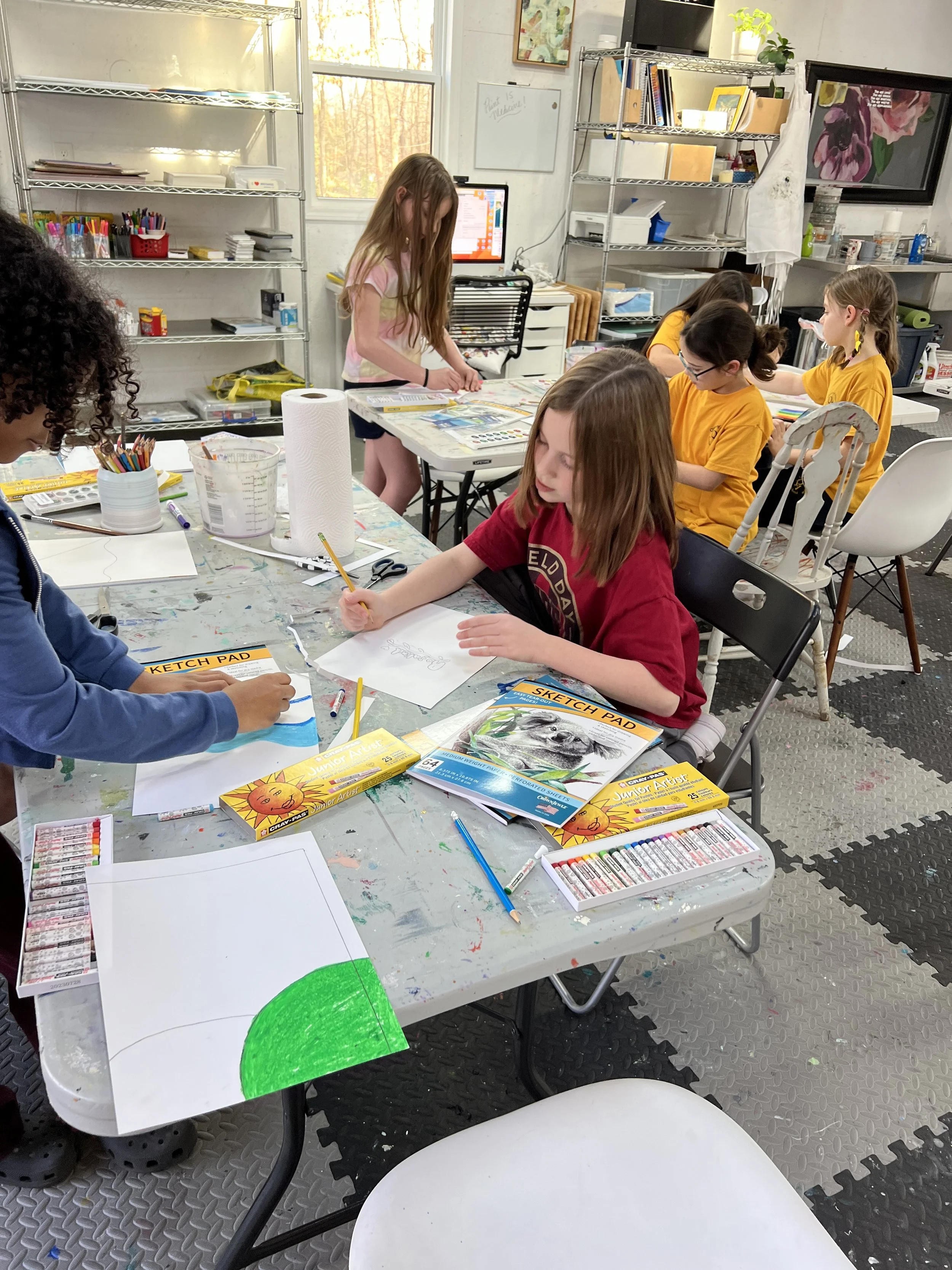 Children in an art classroom working on drawings with colored pencils and markers, with art supplies and papers on the table.