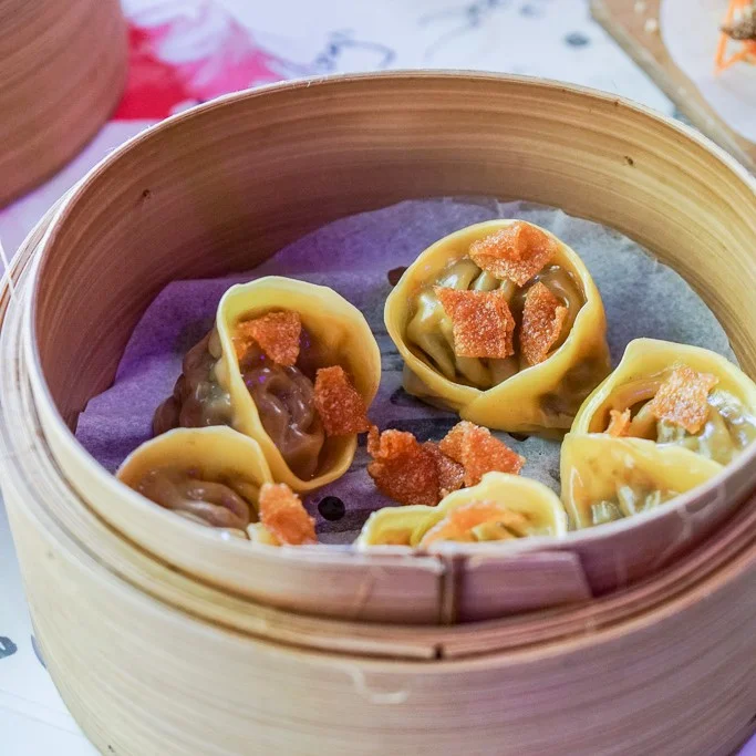 Steamed dumplings with orange topping in a bamboo steaming basket lined with purple paper.