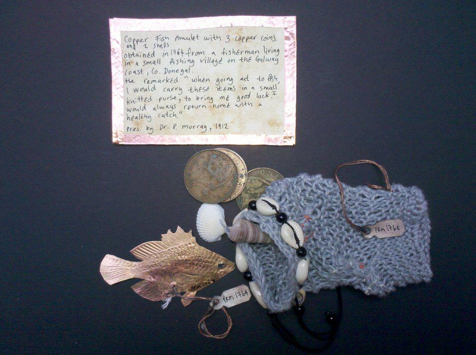 A display of a handwritten note about a copper fish amulet, a small fish-shaped purse, coins, seashells, and a beaded necklace on a dark surface. Community artist working with MIND charity, wellbeing, mental health, Pitt Rivers museum, amulets