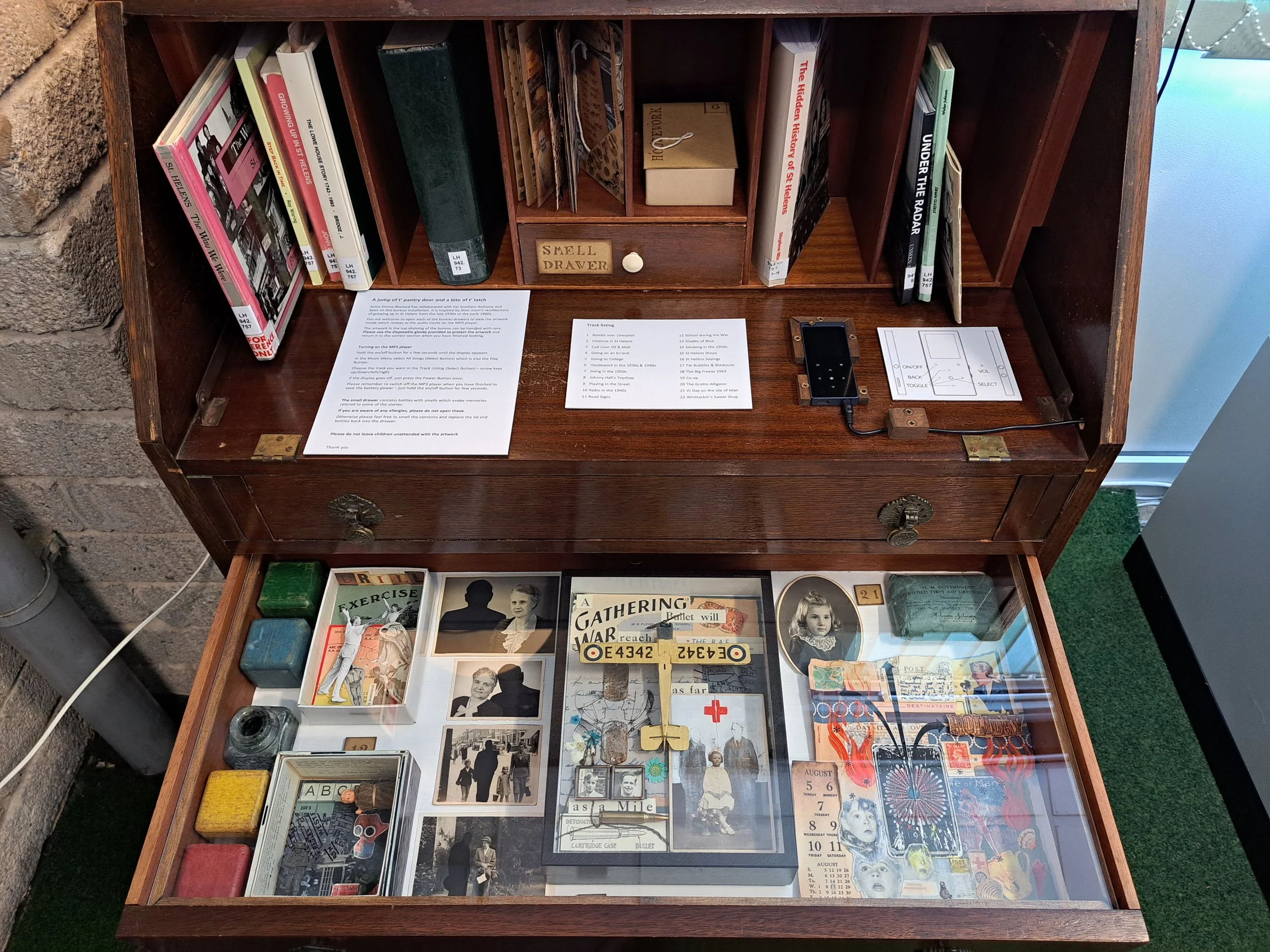 A vintage bureau with books and papers, and a glass display case filled with vintage photographs, tickets, and memorabilia. Artist assemblage art, illustration of life stories, personal diary illustration, collage artist, community artist, nostalgia