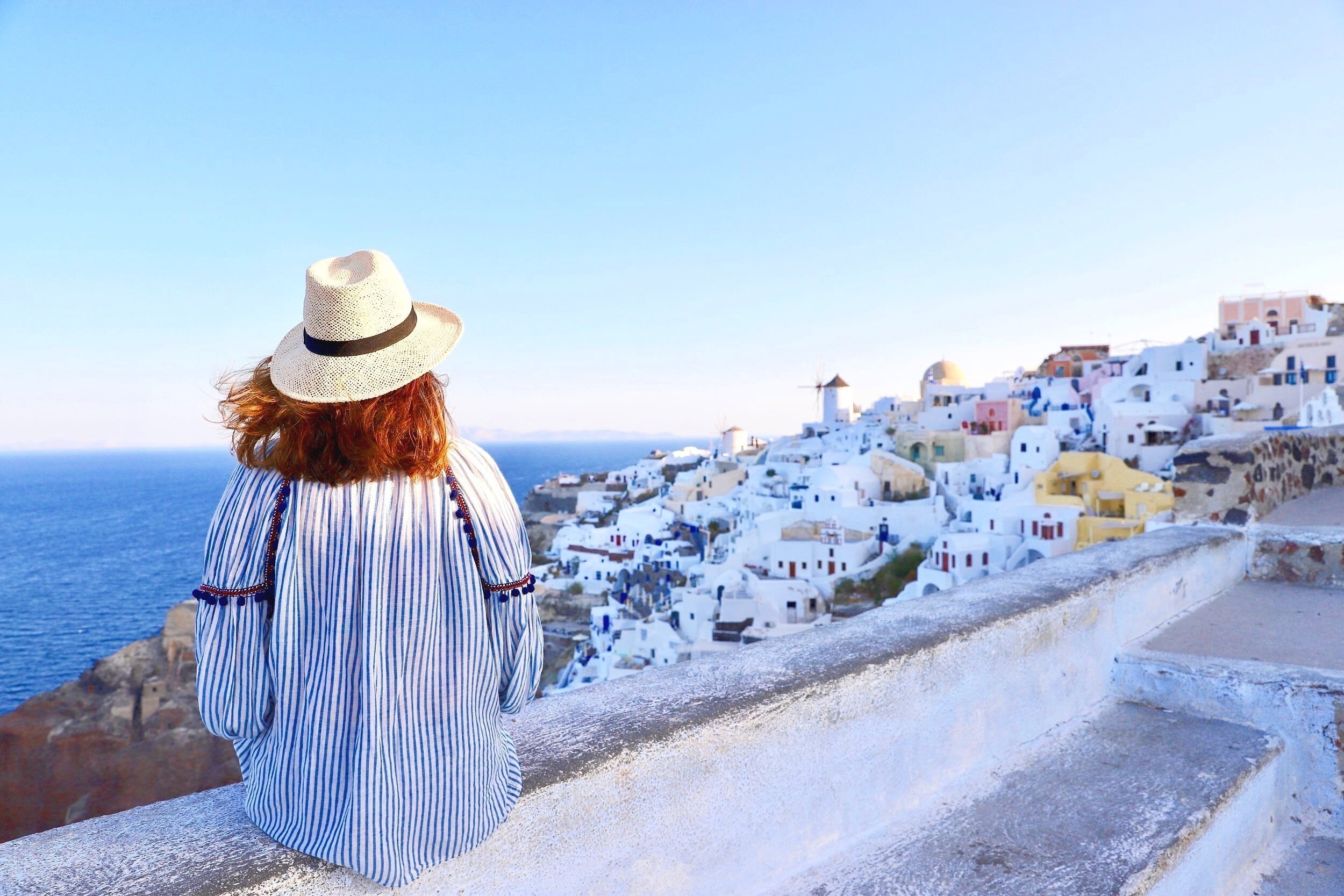 Woman sitting on a stone fence looking at the sea and the village of Oia Santorini. The sky is in blue and pink shade and the woman has a hat on her head and a striped blouse.