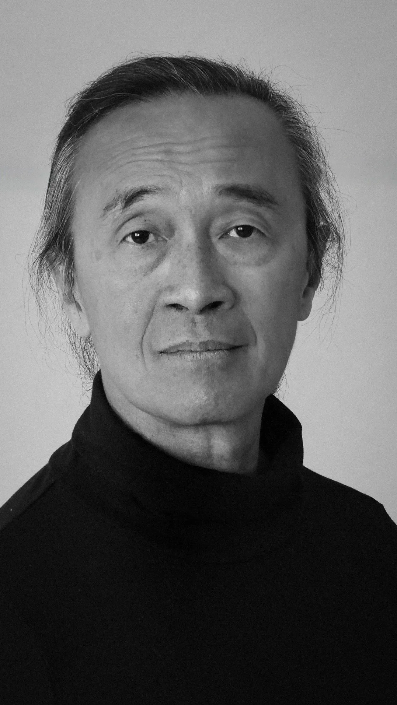 Black and white portrait of an Asian man with slicked-back hair wearing a black turtleneck shirt.