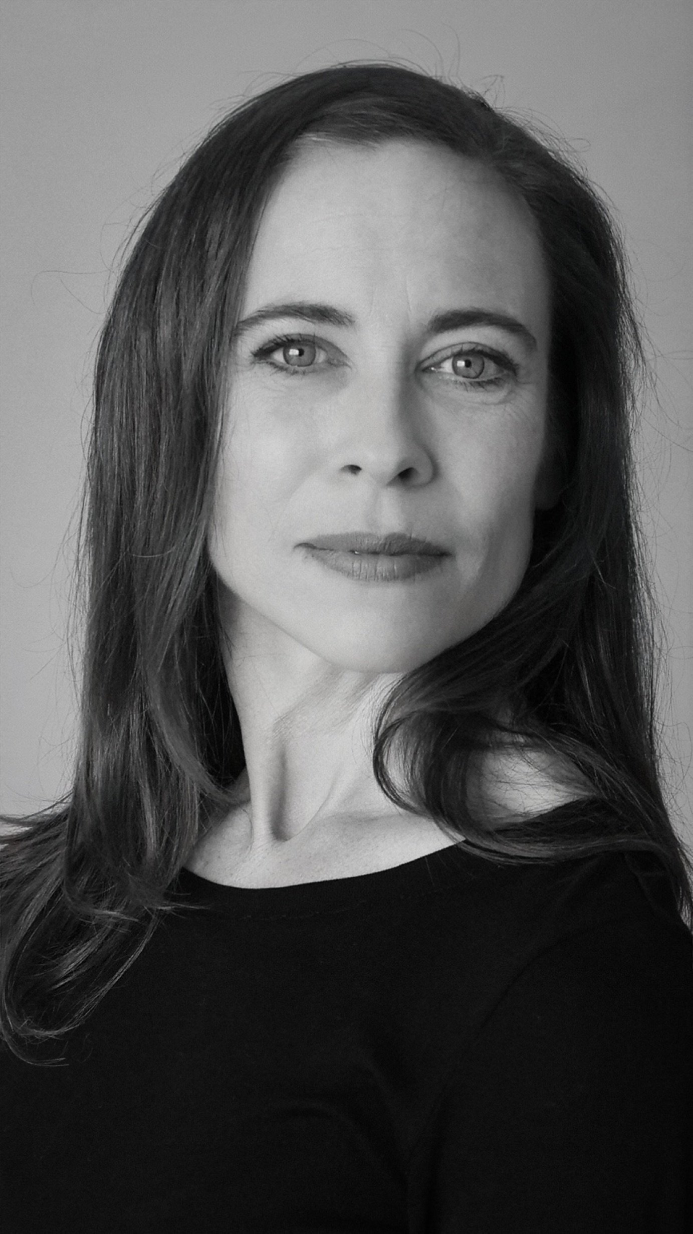 Black and white portrait of a woman with long wavy hair, looking at the camera with a slight smile, wearing a dark top.