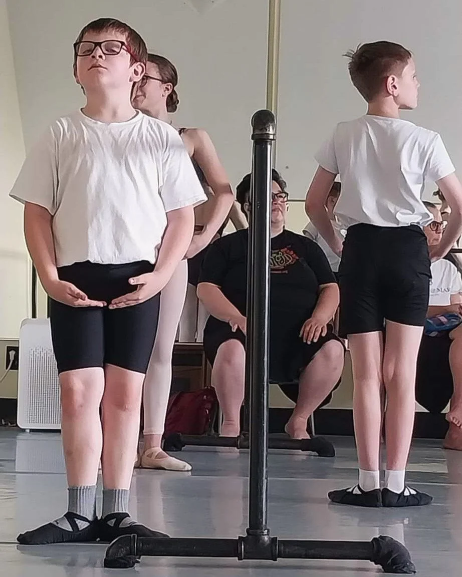 Children in white T-shirts and black shorts practicing ballet at a dance studio, with adults seated and watching in the background.